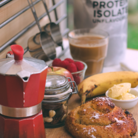 Red espresso maker on a wooden surface with coffee, fruit, and a protein supplement in the background.