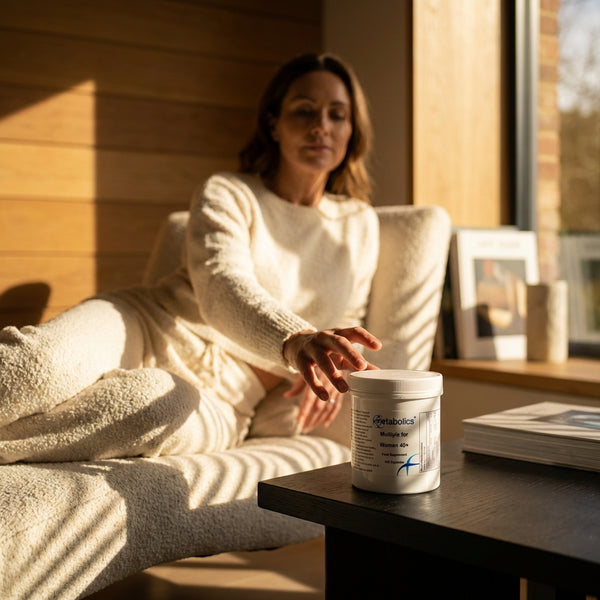 A woman in cream loungewear sits on a cushioned chair, reaching for a tub of Metabolics womens 40+ multiple formula